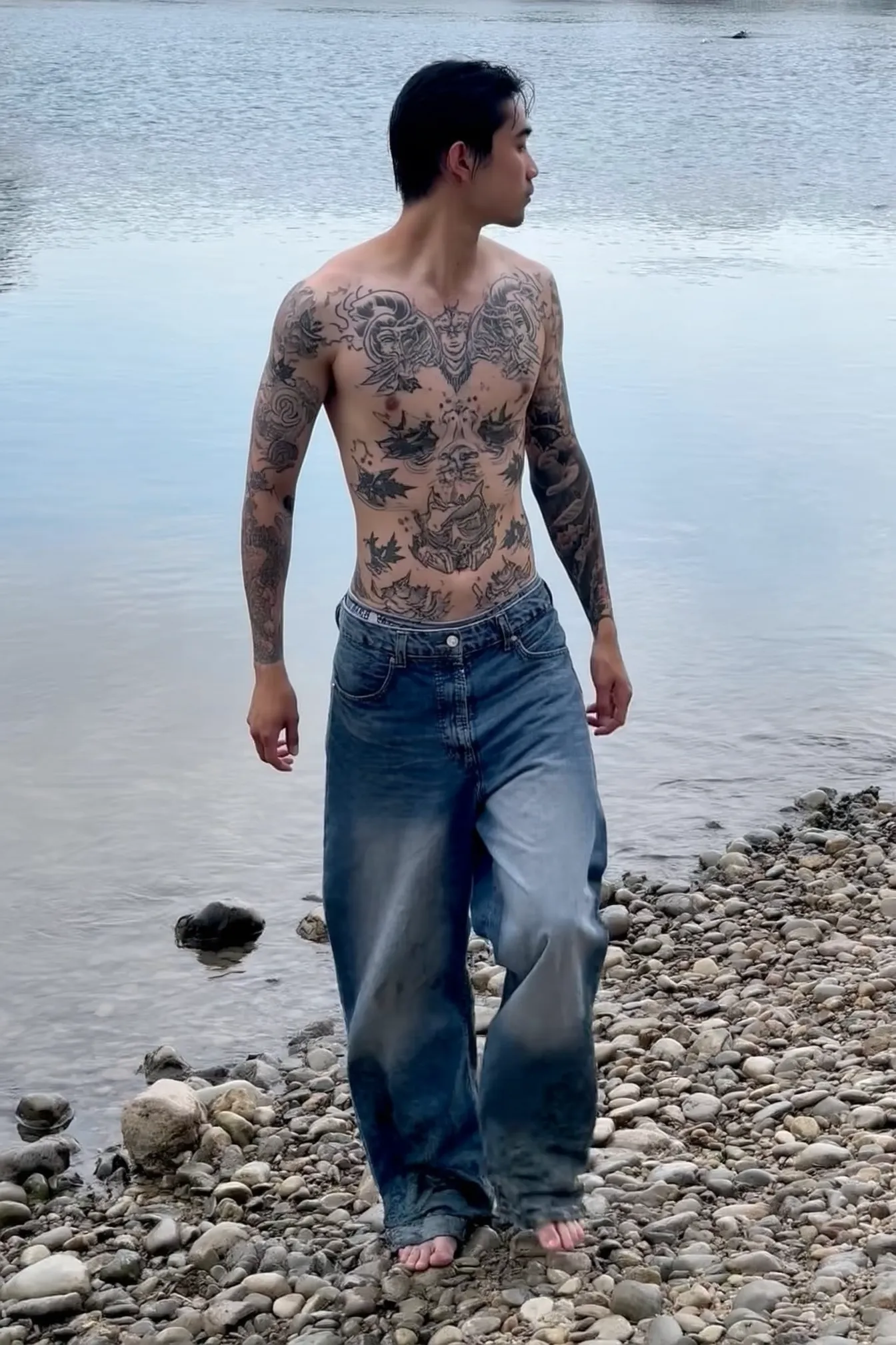 A tattooed man in baggy jeans and no shirt walks barefoot along a rocky riverbank, jeans soaked at the bottom. His torso is fully inked with Hannya masks, waves, and maple leaves. His face is turned downward, arms relaxed. The river reflects pale sky. The colors are muted, skin slightly desaturated in the iPhone camera. Everything feels cool, slow, peaceful — no staging.
