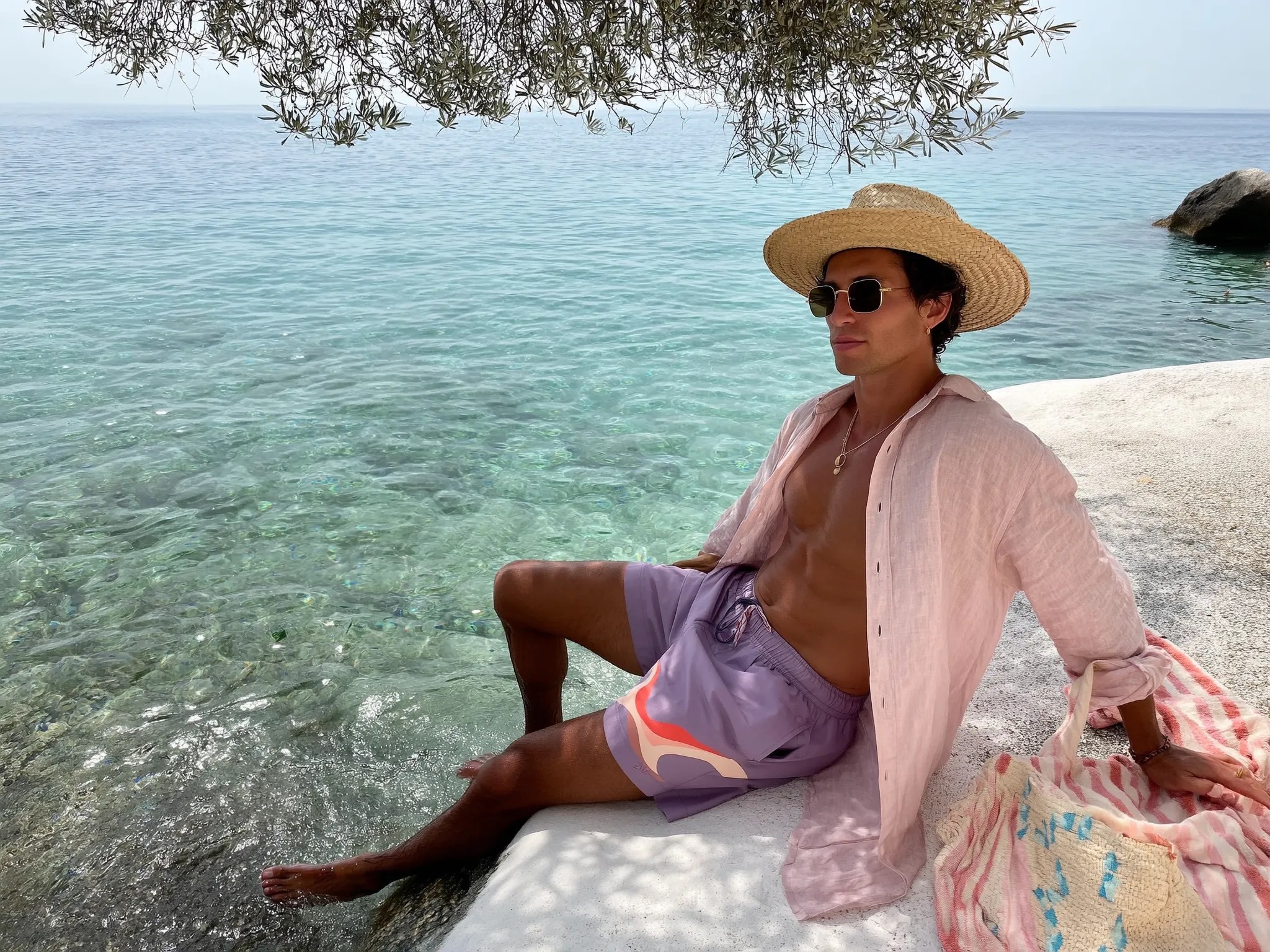 Wide shot taken from about 10 meters away showing a stylish Latina man sitting on sun-warmed, smooth whitewashed stones at the edge of the crystalline Mediterranean sea. He wears tailored swim shorts in a striking dusty lavender with a subtle abstract wavy stripe motif in muted coral and pale peach, crafted from lightweight swim fabric. A loosely draped, unbuttoned blush pink linen shirt adds softness and texture, sleeves rolled casually above the elbow. His sun-kissed skin glows naturally under the soft, clear daylight, complemented by a wide-brimmed boater hat made of woven straw and vintage minimalist gold-rim sunglasses perched slightly down on his nose. He reclines with legs partly submerged in the gently lapping turquoise water, one hand resting on a textured, handwoven canvas tote bag featuring delicate terracotta and sky-blue geometric embroidery. Nearby, a striped pastel towel lies draped over the rocks, while the shimmering sea dominates the foreground and background, reflecting scattered olive tree shadows above. The candid, three-quarter iPhone angle captures tactile textures—wet stone, linen, bronzed skin—and the serene, quietly stylish atmosphere of Mediterranean luxury summer leisure. —hyper-real texture fidelity, natural skin