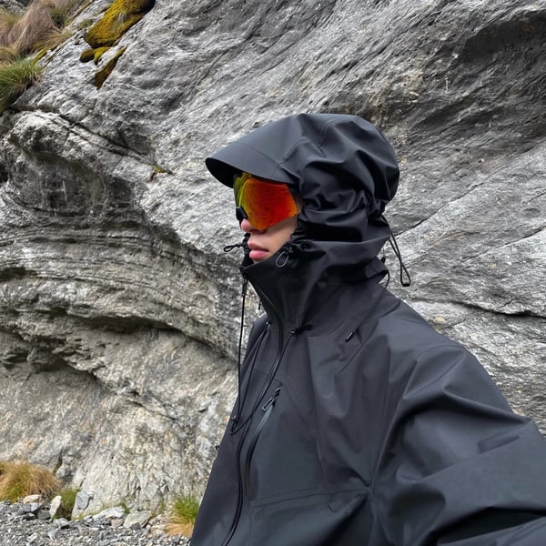 A naturally posed side profile of an individual standing relaxed in front of a textured rugged rock wall within a mountainous terrain. The person wears a matte black technical waterproof hardshell jacket with an oversized hood fully raised, featuring angular shaping and cinch cords creating a sculptural, structured form that closely envelops the head. The jacket shows taped seams and waterproof zippers, highlighting its high-performance, utilitarian design. The individual sports high-contrast, orange-tinted wraparound goggles that vividly reflect the surrounding natural alpine landscape, partially obscuring the face so only the nose and part of the lips are visible beneath the hood and eyewear. Soft, diffused, indirect daylight gently illuminates the scene, emphasizing the rich textures of the waterproof fabric and the coarse rocky surfaces behind. The casual, spontaneous framing includes a subtle, slightly tilted angle, reinforcing the authentic candid iPhone photography aesthetic. This hyper-realistic, cinematic image conveys a calm, tactical, futuristic outdoor vibe within a rugged mountain setting, seamlessly blending minimalist mountain utility with cyberhiker fashion.