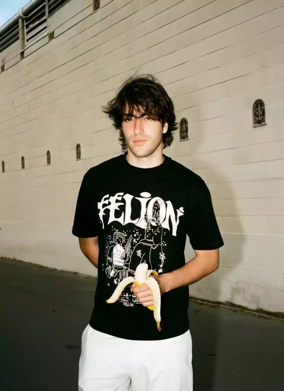 A young man in a black graphic t-shirt and white pants peels and eats a banana, captured in a hyperrealistic photo style suitable for an ai video generator or ai avatar generator in fashion-focused digital art creation.