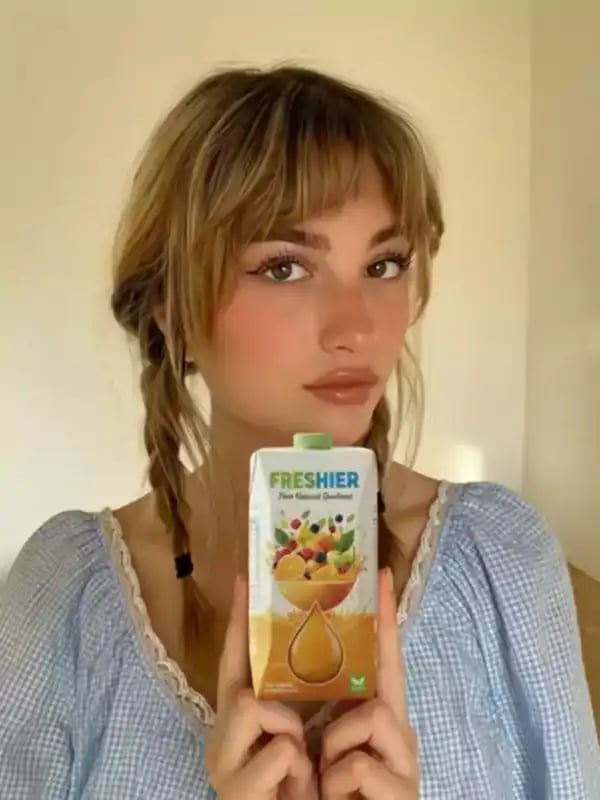 A young woman with braided hair and a blue dress holds and showcases a carton of Freshier fruit juice, demonstrating product placement in a short AI-generated promotional video.