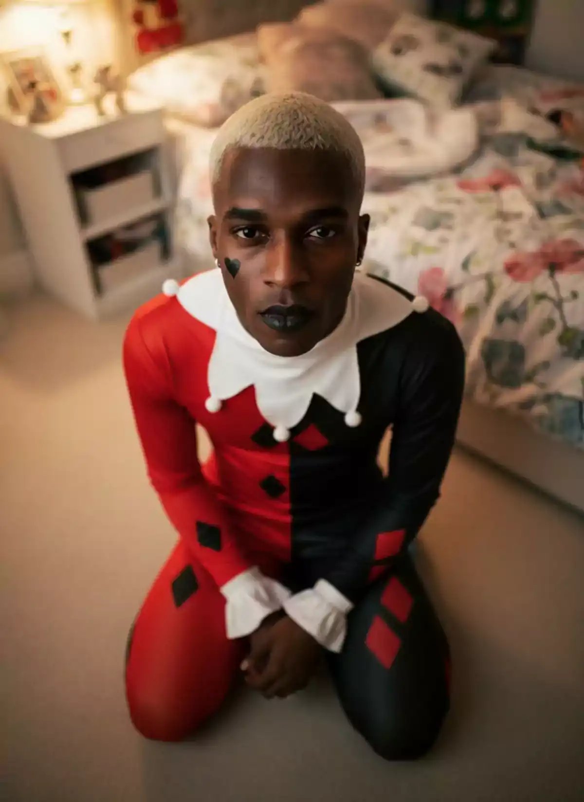 A person wearing a red and black jester costume with a white collar sticks out their tongue while kneeling on a bedroom floor, showcasing hyperrealistic photo style suitable for ai video generator and ai image generator applications.