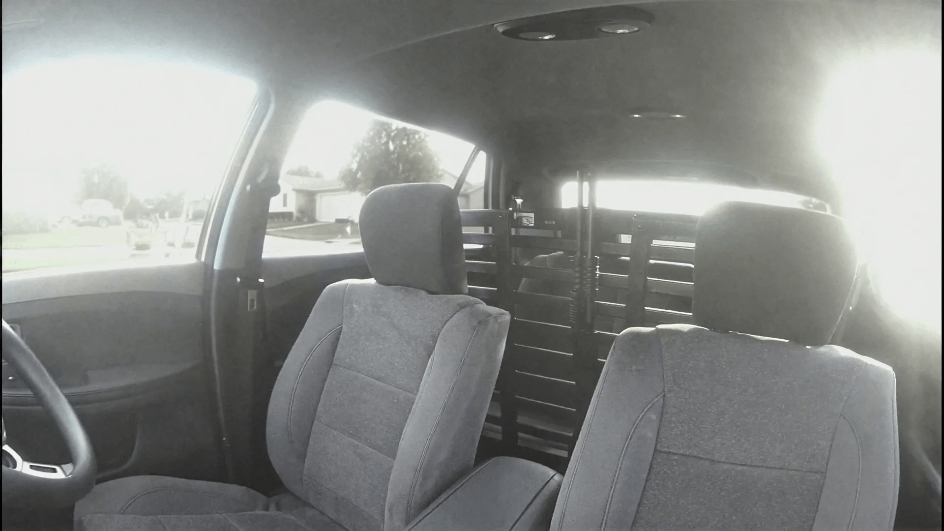 Police cruiser interior, static wide shot from the dashboard facing the passenger seats, empty front seats with gray fabric upholstery, metal police partition cage behind the seats, a shotgun mounted vertically in the center of the partition, bright midday sunlight blasting through the windshield creating harsh overexposed highlights and lens artifacts, suburban houses visible through the windows, slightly washed-out colors, flat digital sensor look, DVR/security camera aesthetic, cheap wide-angle lens distortion, mild compression artifacts, low dynamic range, subtle digital noise, slightly blown highlights, surveillance style framing, raw ungraded footage.