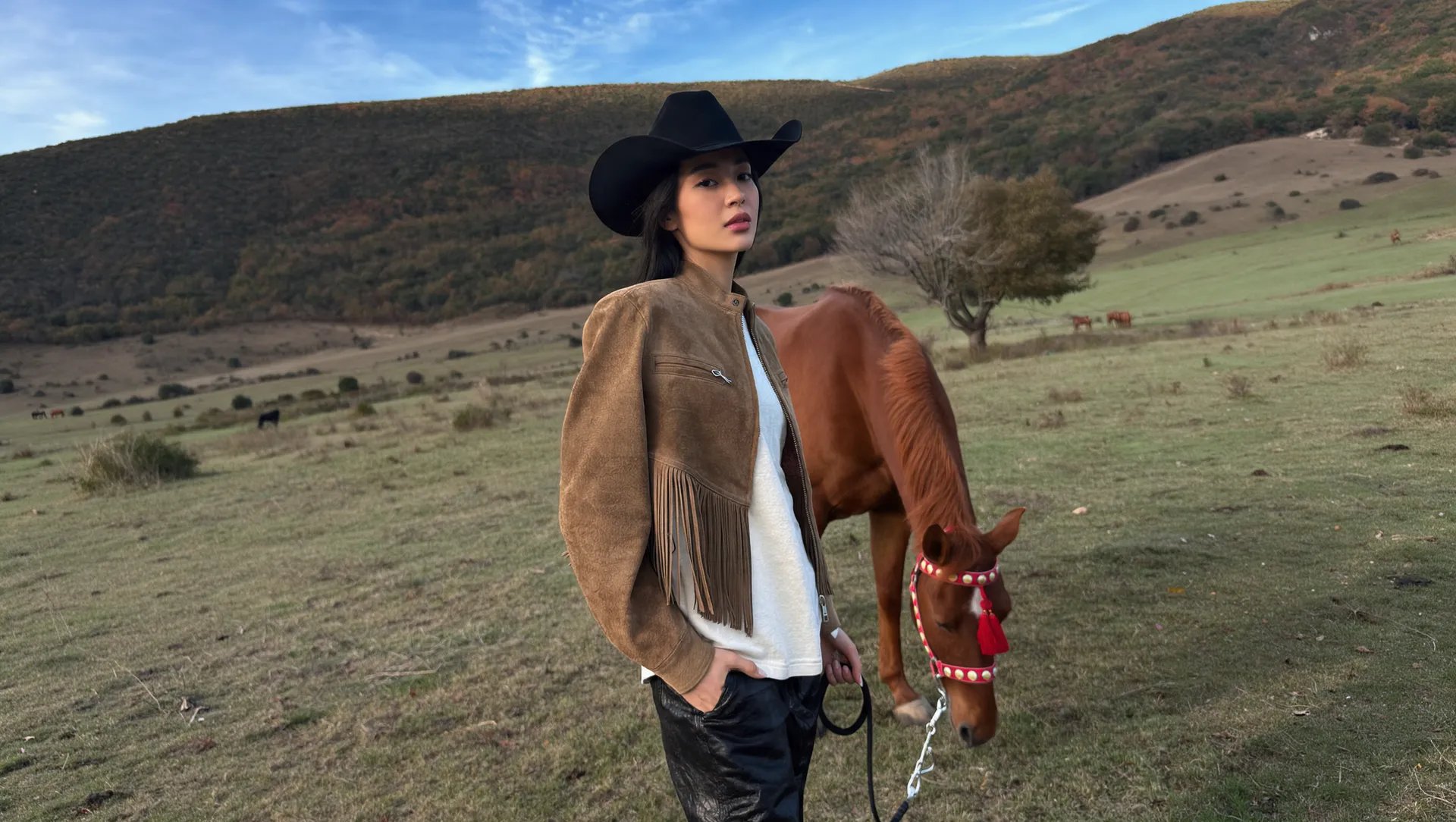 Candid phone-camera look, soft late-afternoon light: Asian woman with long dark hair in a black felt cowboy hat, tan suede jacket with fringe along the sleeves, white tee, black leather trousers, one hand in her pocket, holding the lead of a chestnut horse grazing beside her. Red halter with white studs and red tassels. Wide grassy valley, autumn hillsides, a bare tree and distant horses, pale blue sky with wispy clouds