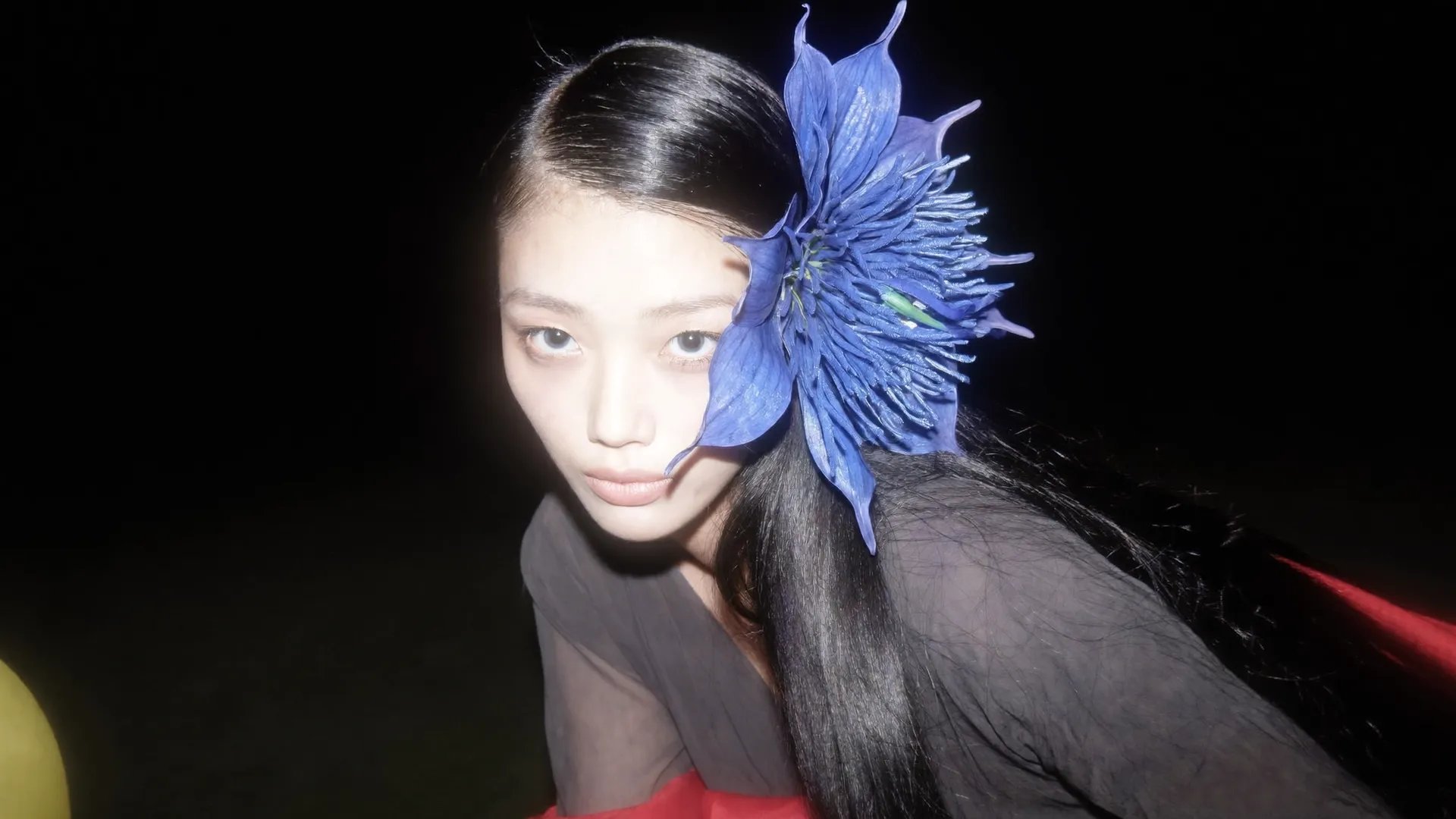 Night portrait, direct on-camera flash, pitch-black background: young Asian woman with long slicked-back black hair leans toward the lens, eyes wide, bare-faced. Oversized blue fabric flower with a fringed center tucked behind her ear. Sheer dark grey top, sliver of red fabric at the frame edge, hard flash falloff, slight grain