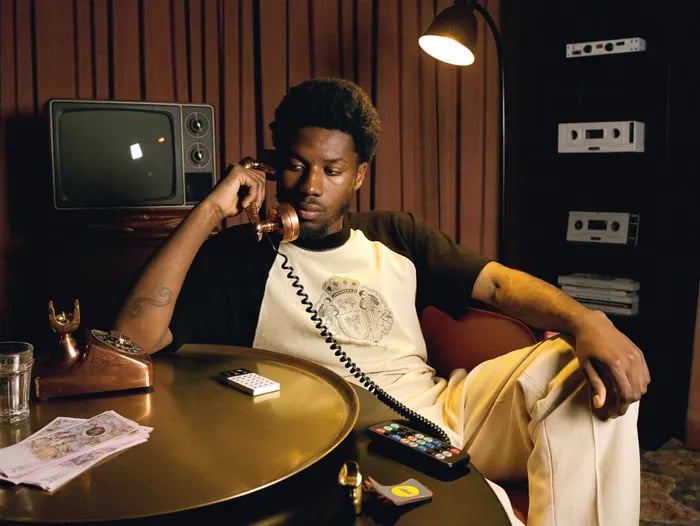 0s portrait, warm tungsten film look: young Black man with a short afro sits in a dark wood-paneled room holding a copper rotary-phone handset to his ear, coiled cord, gaze lowered. Cream raglan tee with brown sleeves and a heraldic crest print, cream trousers, forearm tattoo. Round brass table with the copper phone base, a small white calculator and a glass; wood-cased CRT TV and vintage stereo deck behind him, red armchair