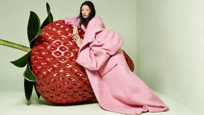 Surreal fashion campaign, soft even studio light: Asian model with long black hair and red lips leans on a hyperreal glossy strawberry as tall as she is, red nails resting on its skin. Enormous fluffy pink mohair coat, oversized sleeves and hem spilling across the floor like a train. Pale sage-green seamless backdrop and floor, deadpan gaze at camera