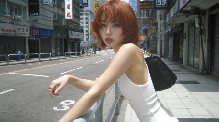 Digicam snapshot, harsh midday sun, slightly blown highlights: young woman with a copper-red shag haircut and bangs looks back over her shoulder, lips parted, arm draped over a metal street railing. White ribbed tank top, black quilted leather bag on a gold chain, silver ring. Empty Hong Kong street behind her: white road markings, grey concrete buildings, red-and-white Chinese signage, shuttered shopfronts