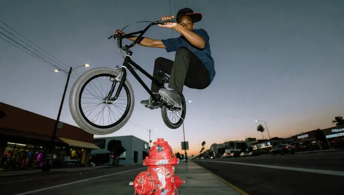 Low-angle street photo at dusk: BMX rider in a navy tee, black pants and dark cap frozen mid-air on a black bike with white tires, bike tilted, jumping over a sticker-covered red fire hydrant in the foreground. Blue-to-orange twilight sky, streetlights just switched on, storefronts and a palm tree along a wide LA boulevard, distant headlights, subject lit with a touch of flash